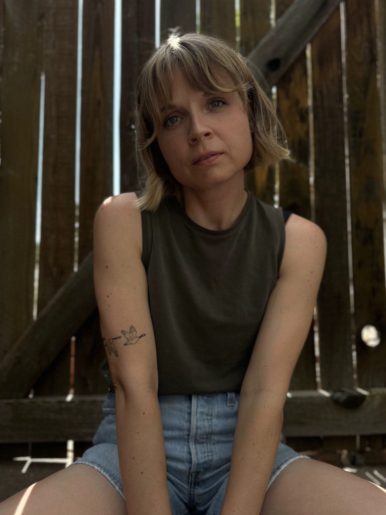 Woman with a blond bob, a green tank top, and light denim shorts sits in front of a fence. She has a tattoo of flying geese on her left arm and a septum pierecing.